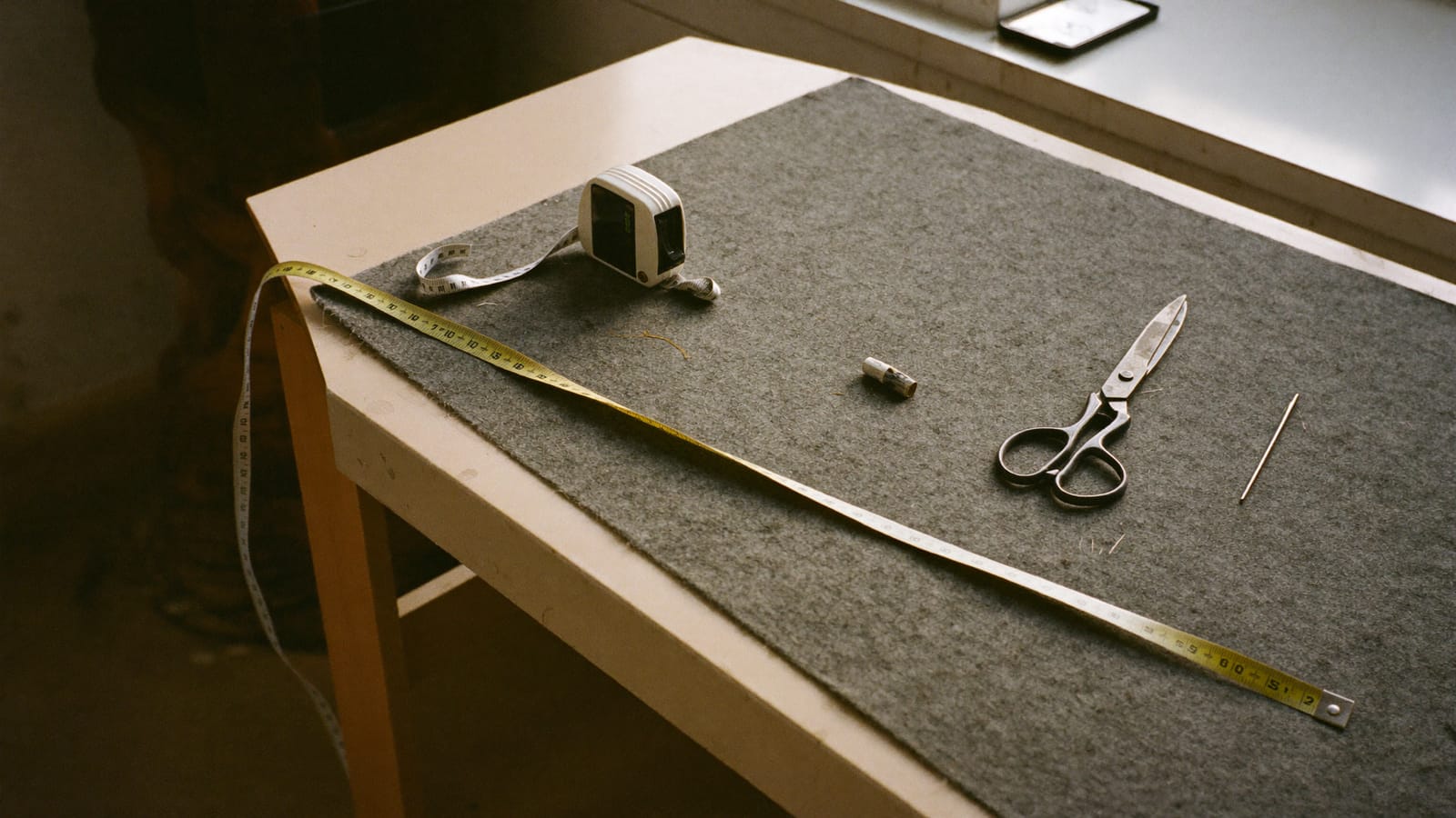 A tape measure, chalk, shears and a pincushion lying on grey wool across a tailor's workbench in afternoon light