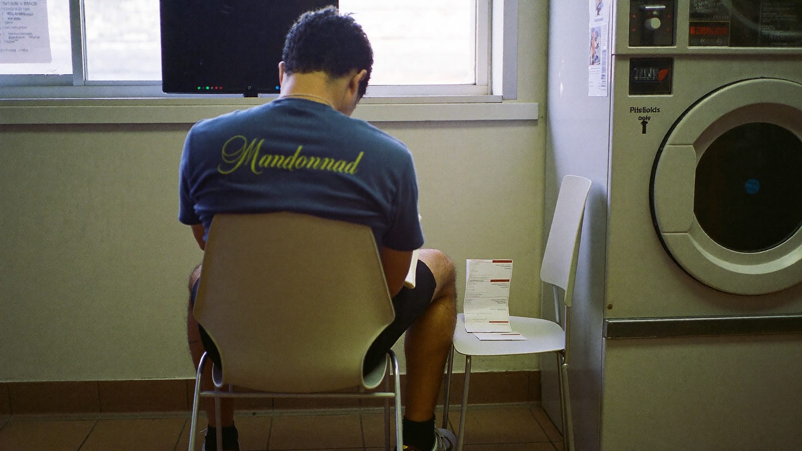 A man seen from behind on a plastic chair in a laundromat, writing in a notebook while a washing machine turns behind him.