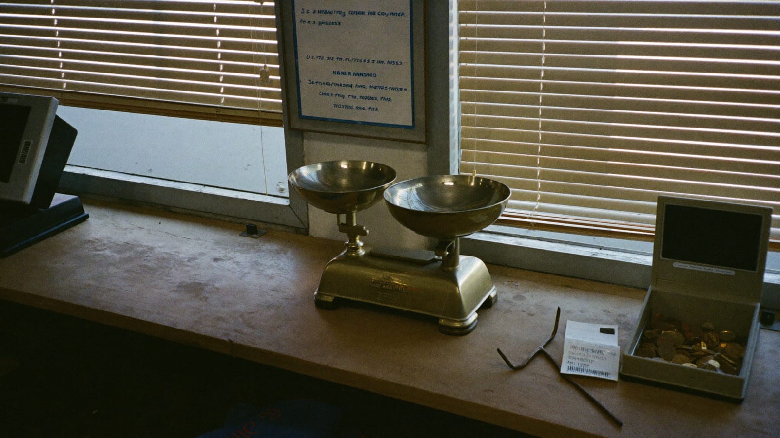 A brass balance scale and a set of weights on a hardware store counter beside a coin tray and a receipt spike at dusk.